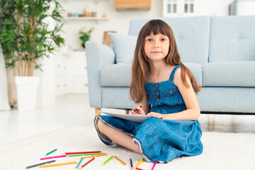 A cute little girl sits on the floor at home and draws with colored pencils. Child development and creativity for preschoolers and younger students