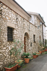 An old Italian stone house with iron bars on the windows and an arched door. Near the facade, there are planters with trees and flowers.