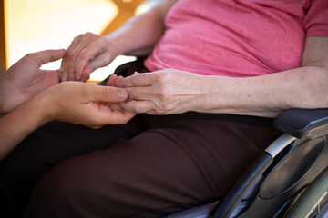 gros plan sur les mains d'une personne âgée en fauteuil roulant tenant celles d'un jeune homme....