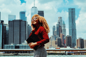 Portrait smiling young adult entrepreneur millennial woman with eyeglasses and afro hair talking on a phone call outdoors with Manhattan New York City skyline skyscraper behind Hudson river - obrazy, fototapety, plakaty