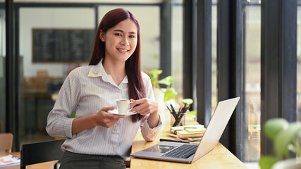 Young asian woman holding cup of coffee in her hands and smiling to camera, sitting in coffee shop