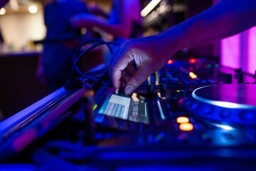 A DJ on a party mixing, controlling the volume and making music on stage with DJ equipment during the night in a colourful setting.