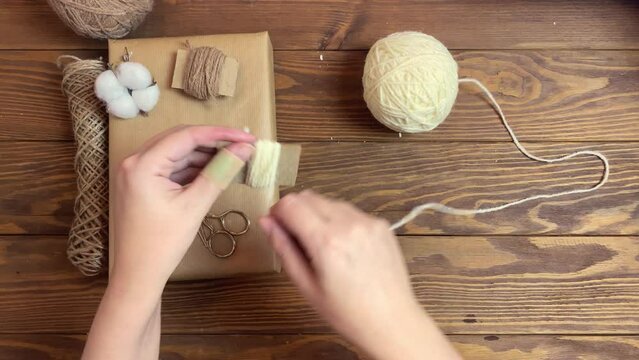 Girl's hands wind white yarn on piece of cardboard. Box in craft wrapper, twine, cotton and scissors on wooden table. Festive mood. Handmade crafting decorations of gift for Christmas or Birthday.