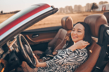 Happy smiling brunette woman driver sitting in new red cabriolet car on beach coast, smiling looking at camera enjoying journey. Driving courses and life insurance concept. Glamorous lifestyle