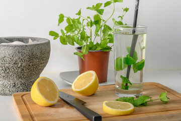 Moroccan mint in a pot, and in a glass, a sliced lemon and a black knife on a chopping board. Hot water in the glass. Cleanse, and a healthy lifestyle. There is a granite mortar in background