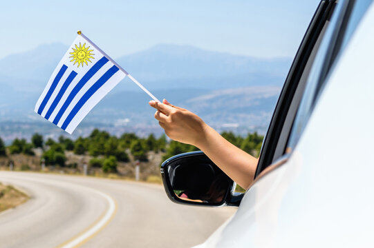 Woman holding Uruguay flag from the open car window driving along the serpentine road in the mountains. Concept