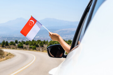 Woman holding Singapore flag from the open car window driving along the serpentine road in the mountains. Concept - 517346996