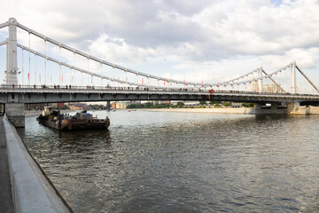 Moscow river with barge and cargo, pedestrian bridge with pedestrians. Summer day, cloudy sky. Center of Moscow.