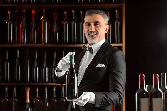 Elegant waiter sommelier wine steward with a bottle of wine. Examination of wine products. Restaurant staff among shelves of wine bottles. Stylish middle-aged man with a grey beard.