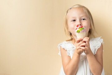 Beautiful little caucasian girl with blonde hair in a white dress enjoying berry flavored waffle cone ice cream on the beige background