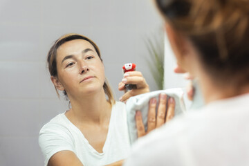 Reflection of young woman wiping the mirror while spraying cleaning agent on the glass