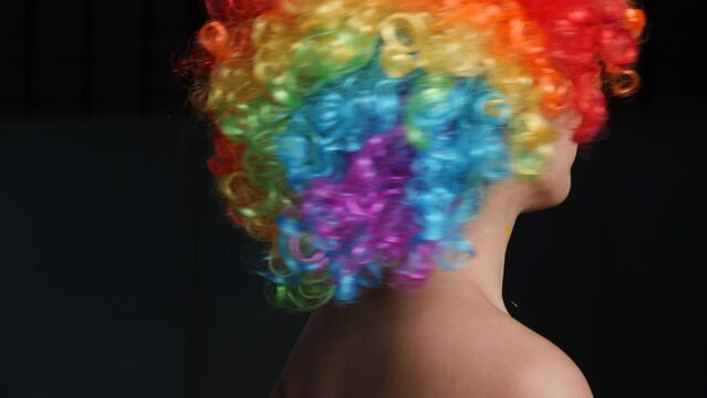 Close-up portrait. Smiling young attractive female in rainbow wig with paint on her cheeks on black background, slow motion. Concept of pride