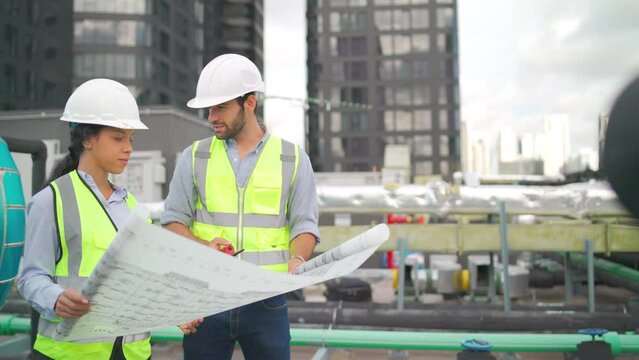 Side and front view of two professional engineer or workers hold building plan or blue print of construction site and they stay in area of rooftop of the building.