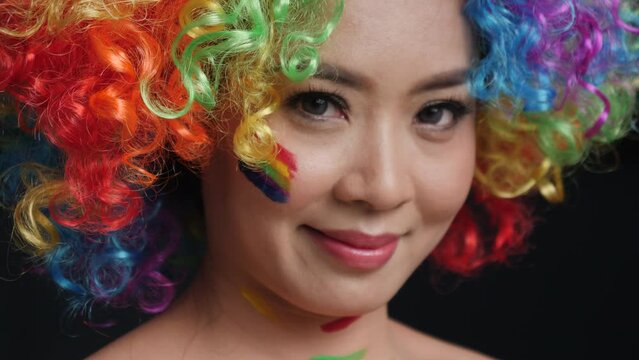 Close-up portrait. Smiling young attractive female in rainbow wig with paint on her cheeks on black background, slow motion. Concept of pride