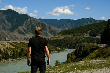 A young man in a black T-shirt enjoys the view of the mountains and the river. Altai, Russia.