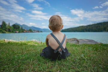 Fototapeta premium back of a blond german baby boy in bavarian dress with lederhose sitting on the meadow by the shore of the famous bavarian lake Walchensee with turquoise blue water in the background and watching 
