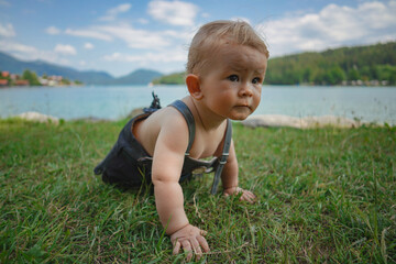 Naklejka na ściany i meble cute baby boy in bavarian dress with lederhose is crawling on the meadow by the shore of the famous bavarian lake Walchensee with turquoise blue water in the backround