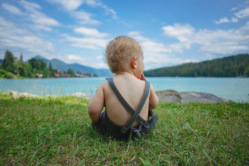 Fototapeta premium back of a blond german baby boy in bavarian dress with lederhose sitting on the meadow by the shore of the famous bavarian lake Walchensee with turquoise blue water in the background and watching 