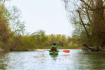 Rear view of young woman in green kayak paddle at river near trees with gentle green leaves at spring