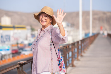 Happy mature female traveler with hat waving hand looking at camera while boarding the ferry in...