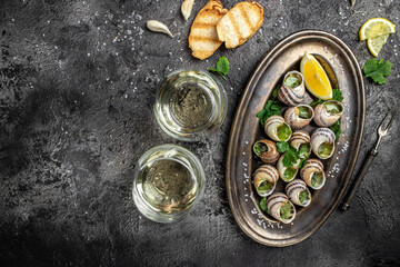 Delicious cooked sea escargo snails with herbs, butter, garlic on metal plate with forks. wine glass. gourmet food. Restaurant menu, Traditional French cuisine