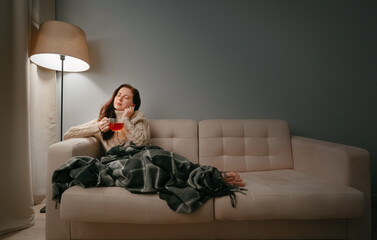 Woman with a cup of cough tea sits in a blanket on the sofa at home. The concept of the common cold.