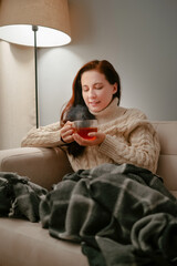 Woman sits on a sofa with a large mug of fragrant healing tea on a winter evening. Influenza and viruses concept.