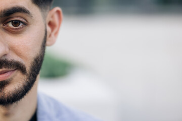 Half face of upset bearded caucasian young man looking straight to camera while standing outdoors in empty town. Half face portrait of young man with beard