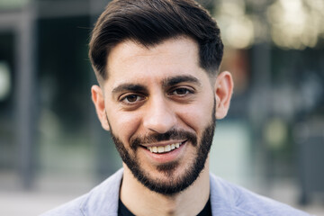 Obraz premium Face portrait of attractive joyful young caucasian mixed race man in business formal suit smiling and posing near modern buildings outside. Businessman. Ethnicity