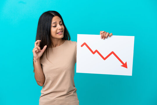 Young woman over isolated background holding a sign with a decreasing statistics arrow symbol with fingers crossing