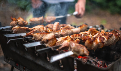 Man hand straightens the skewer with barbecue delicious grilled meat on brazier outdoors. Person preparing pork steaks with smoke during picnic at the nature. Hot BBQ closeup