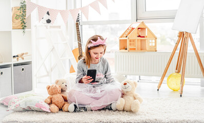 Lovely little girl holding phone sitting on floor with plush teddies