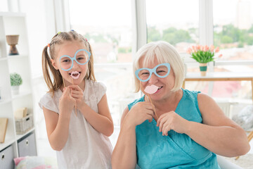 Happy little girl with grandmother putting on glasses and lips on sticks in light room