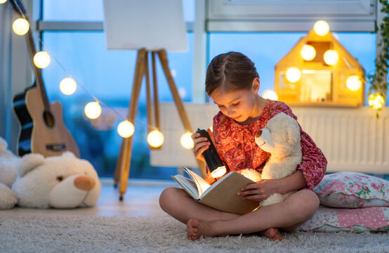 Little girl reading fairy tales book in the evening with lantern before going to sleep. Cute child sits on the floor with Teddy bear and flashlight.