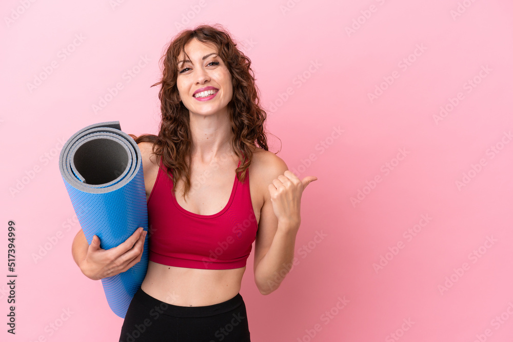 Wall mural Young sport woman going to yoga classes while holding a mat isolated on pink background pointing to the side to present a product