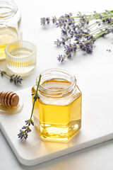 Lavender honey in opened glass jar with  wooden honey dipper, drizzler and lavender flowers on a white board and background.