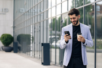 Confident caucasian armenian man using mobile phone holding coffee cup in modern city district. Positive business guy browsing financial news smiling standing outdoor. Successful adult man