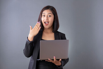 Surprised Mexican woman holding a laptop with a black blazer on a gray background