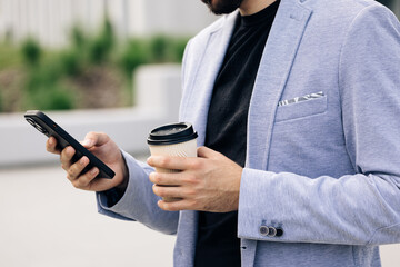 Attractive bearded handsome man with nice hairstyle and beard using his smart phone and drinking coffee typing messages. Looking very stylish and gorgeous