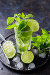 Mojito cocktail, lime slices, mint and ice on a dark background. Summer refreshing cocktail, ice tongs and cocktail spoon on a black plate