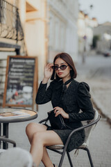 a brunette girl in a short dark dress and black glasses sits on a chair in a cafe in the city