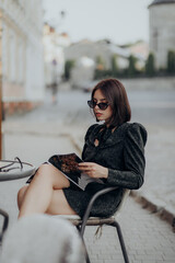 a brunette girl in a short dark dress and black glasses sits on a chair in a cafe in the city