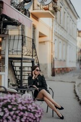 a brunette girl in a short dark dress and black glasses sits on a chair in a cafe in the city