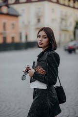 a brunette girl in a short dark dress and black glasses walks down the road in the city with a magazine
