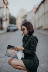 a brunette girl in a short dark dress and black glasses sits on the road in the city with a magazine