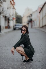a brunette girl in a short dark dress and black glasses sits on the road in the city