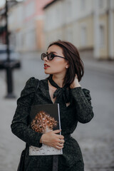 a brunette girl in a short dark dress and black glasses walks down the road in the city with a magazine