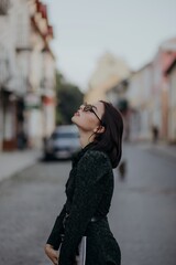 a brunette girl in a short dark dress and black glasses walks down the road in the city with a magazine