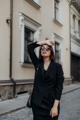 a brunette girl in a black jacket and dress and black glasses walks down the street in the city with a handbag