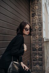 a brunette girl in a black jacket and dress and black glasses walks down the street in the city with a handbag